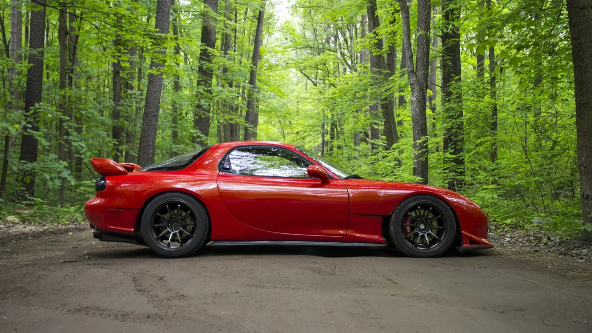 Side profile of a red third-generation Mazda RX-7 FD parked on a dirt road through a dense green forest, lowered on dark multi-spoke wheels with its distinctive rear spoiler.