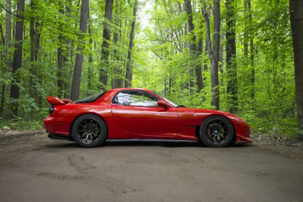 Side profile of a red third-generation Mazda RX-7 FD parked on a dirt road through a dense green forest, lowered on dark multi-spoke wheels with its distinctive rear spoiler.